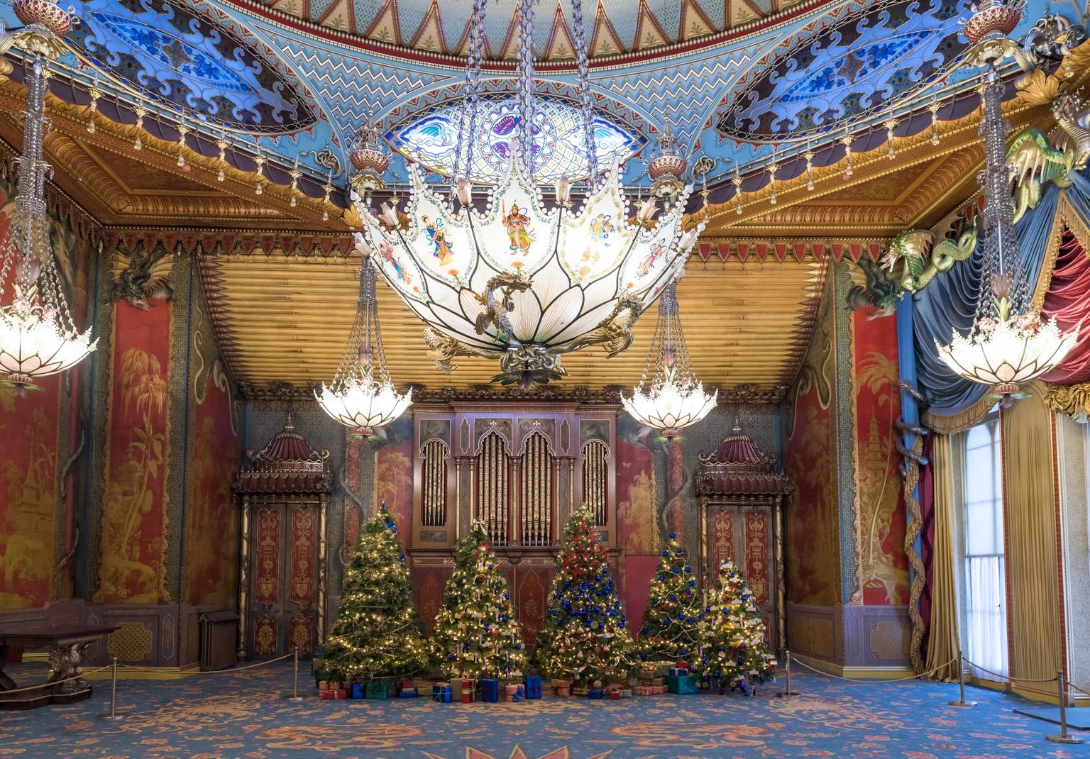 Lavishly decorated music room with stained-glass chandeliers hang above a row of lit Christmas trees and wrapped presents in front of a grand organ.