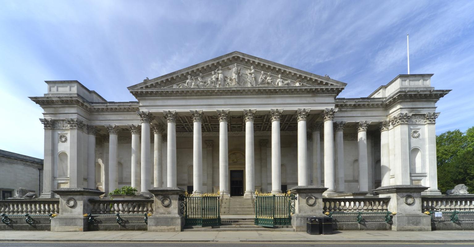 White neoclassical building with tall columns and an ornate pediment stands beneath a clear blue sky.