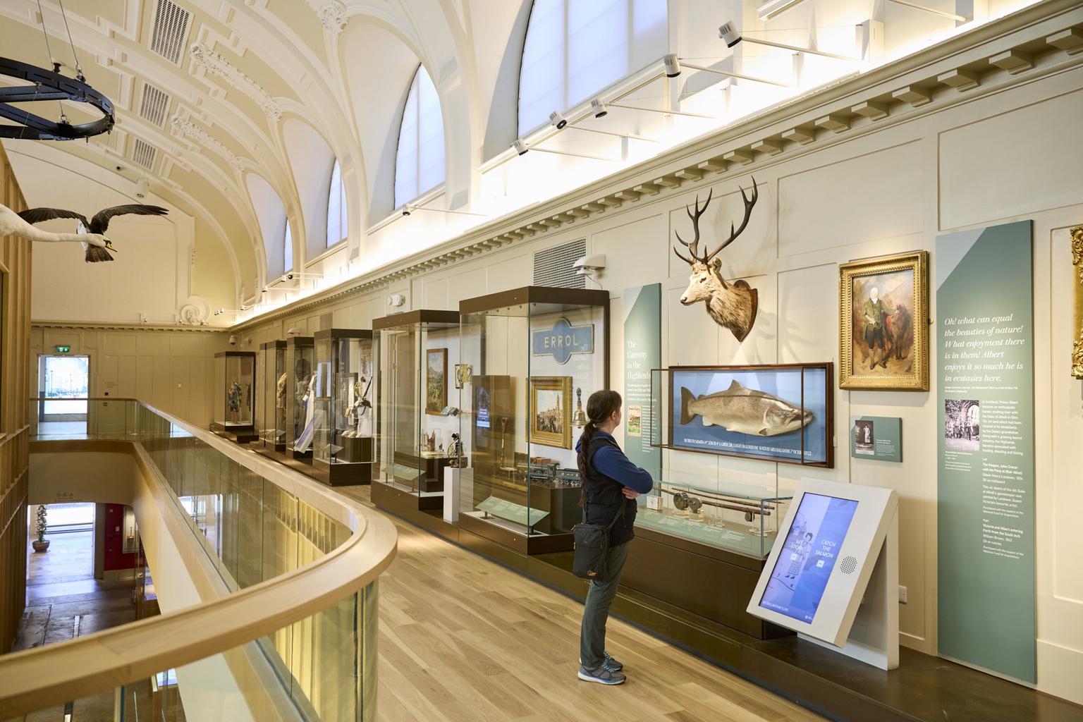 A woman in a museum looks at a large mounted fish, on display below a taxidermy stag head.
