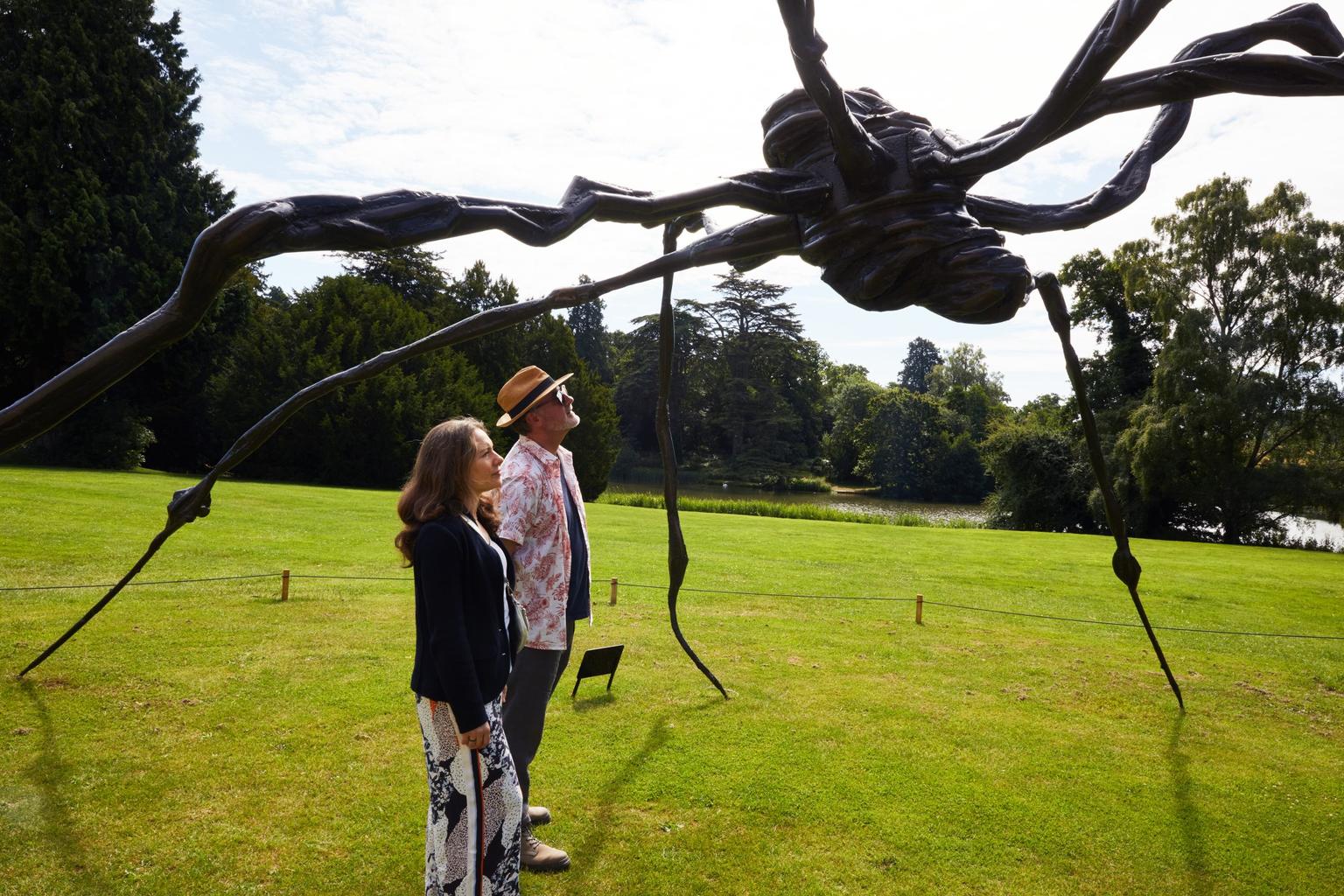 Two people standing on a green lawn, looking up at a towering metal sculpture of a spider.