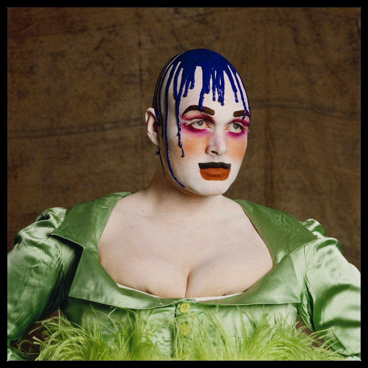 Leigh Bowery poses in dramatic makeup, with bright pink eyeshadow, orange blush, overdrawn red lips with a thick black line, dark block eyebrows and a white base, while blue paint drips from the top of his head. He wears a green satin shirt that reveals his pushed together chest.