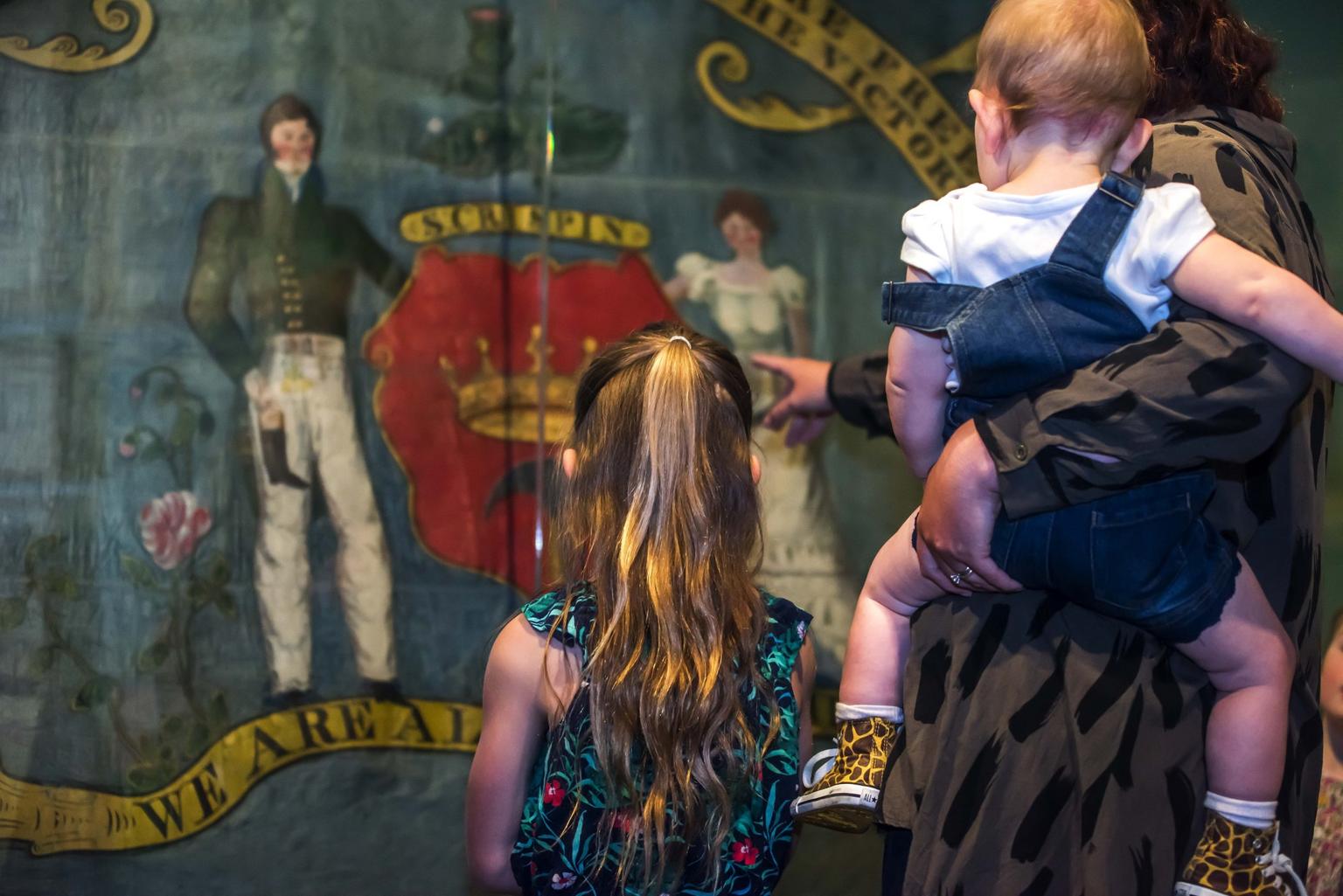 A young girl and woman holding a baby on her hip look into a museum case with a banner in it