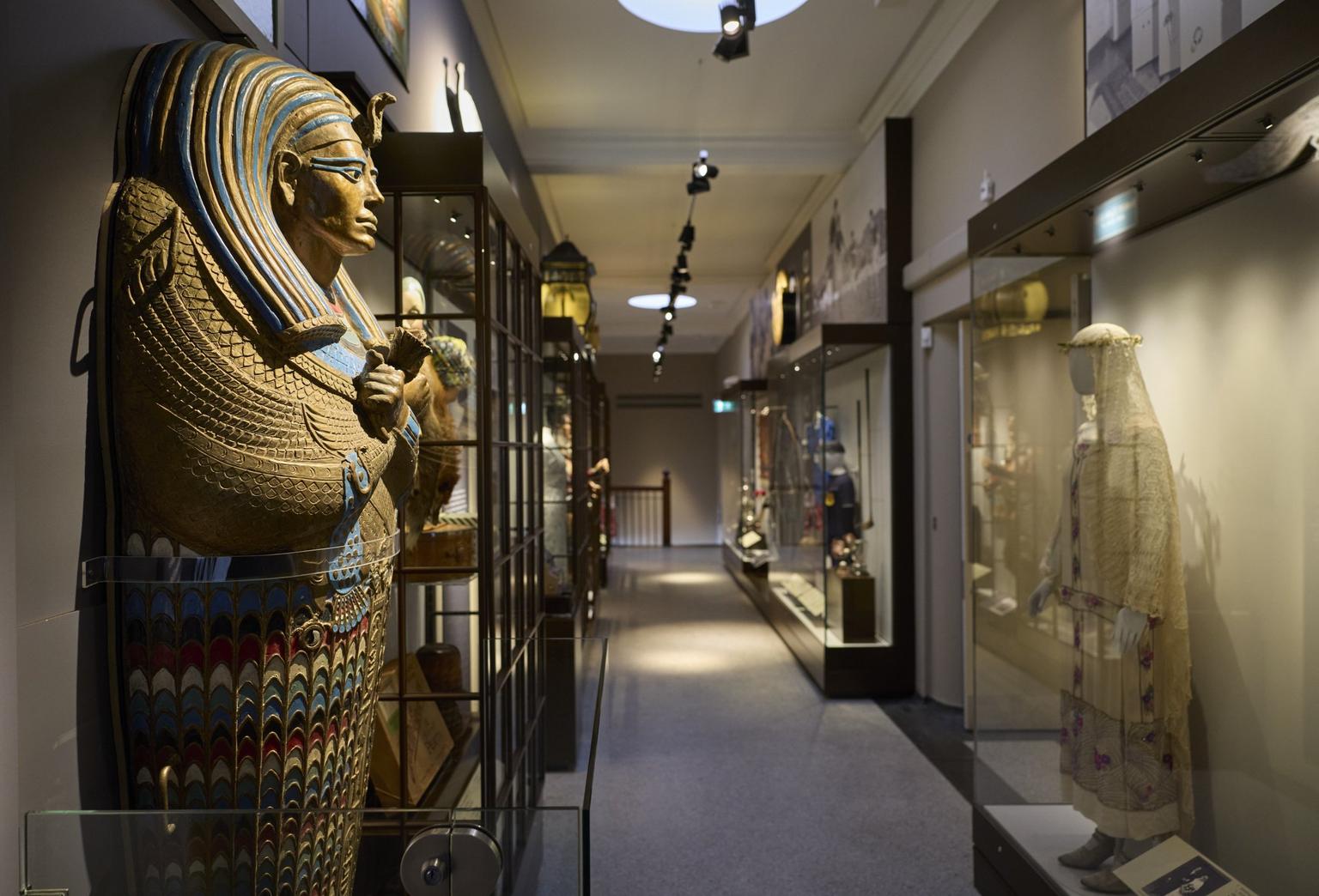 A narrow room with displays of a mummy and historic dress behind glass cabinets.