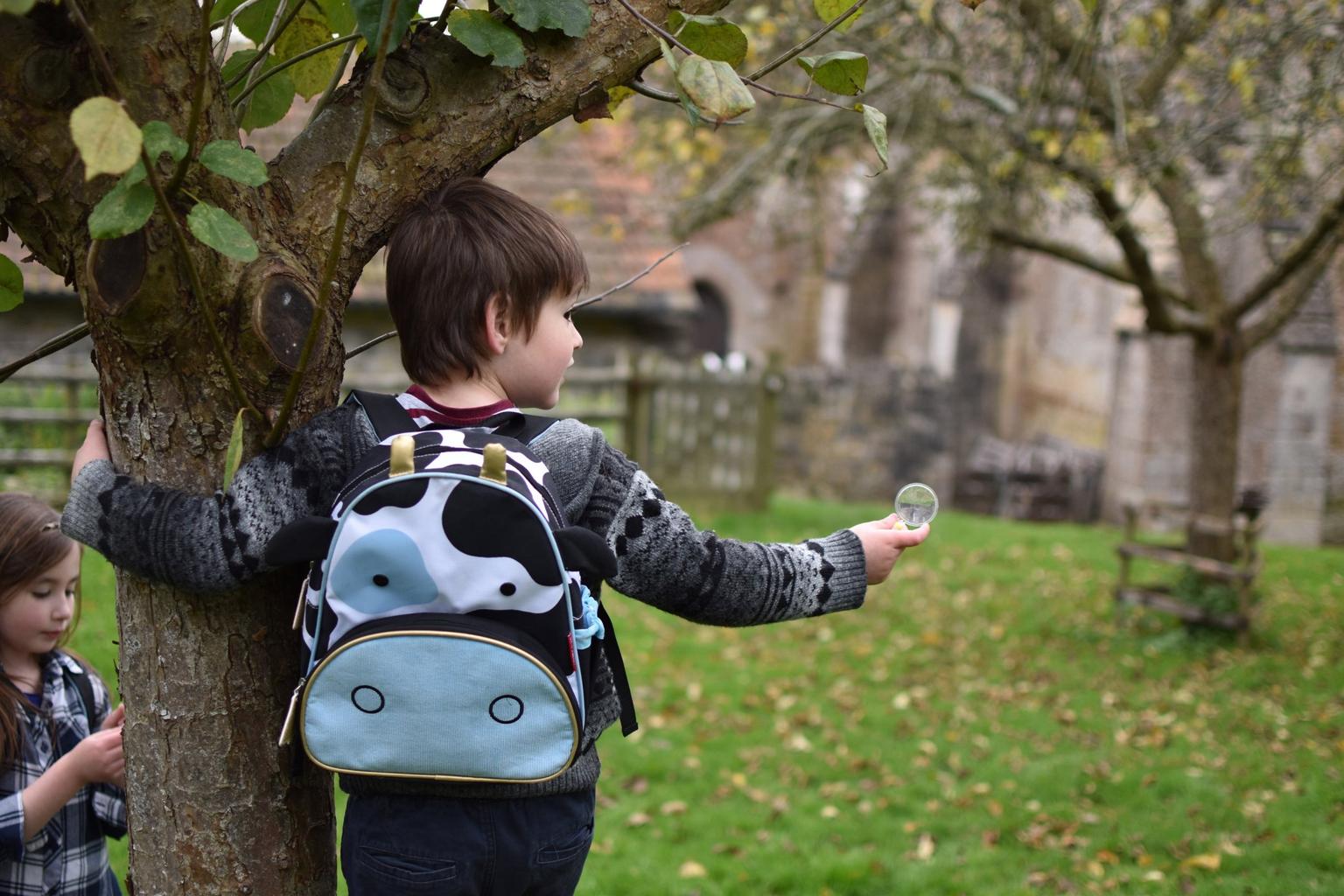 A boy with one arm around a tree, the other hand holding a magnifying glass