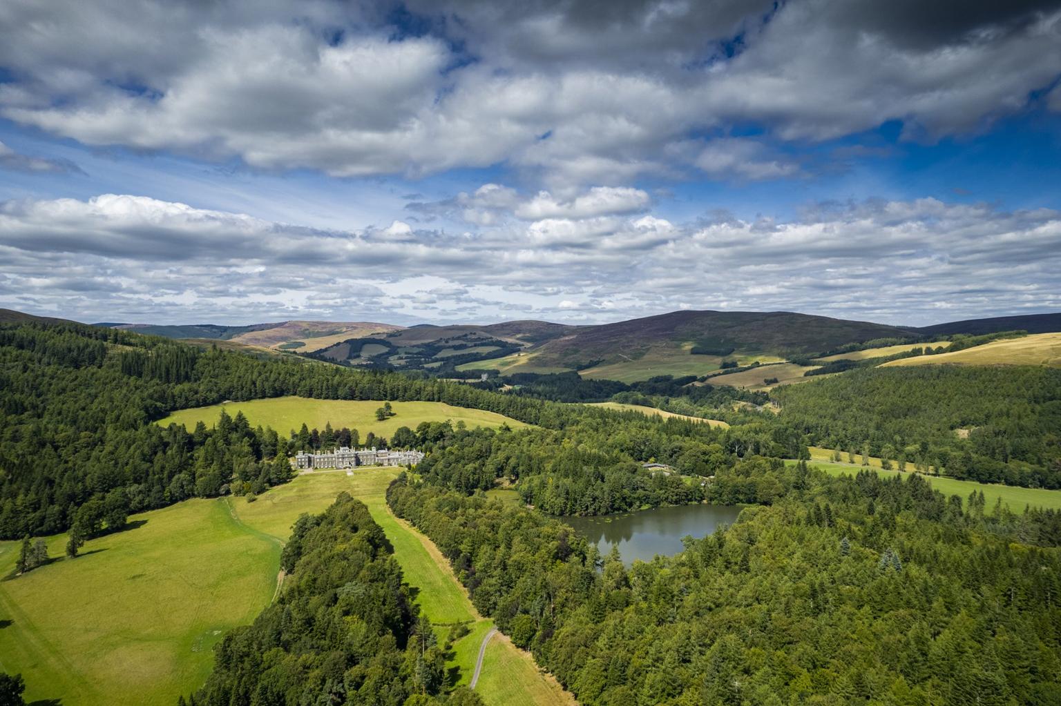 An aerial shot of a stately home in the far distance, surrounded by forests and hills