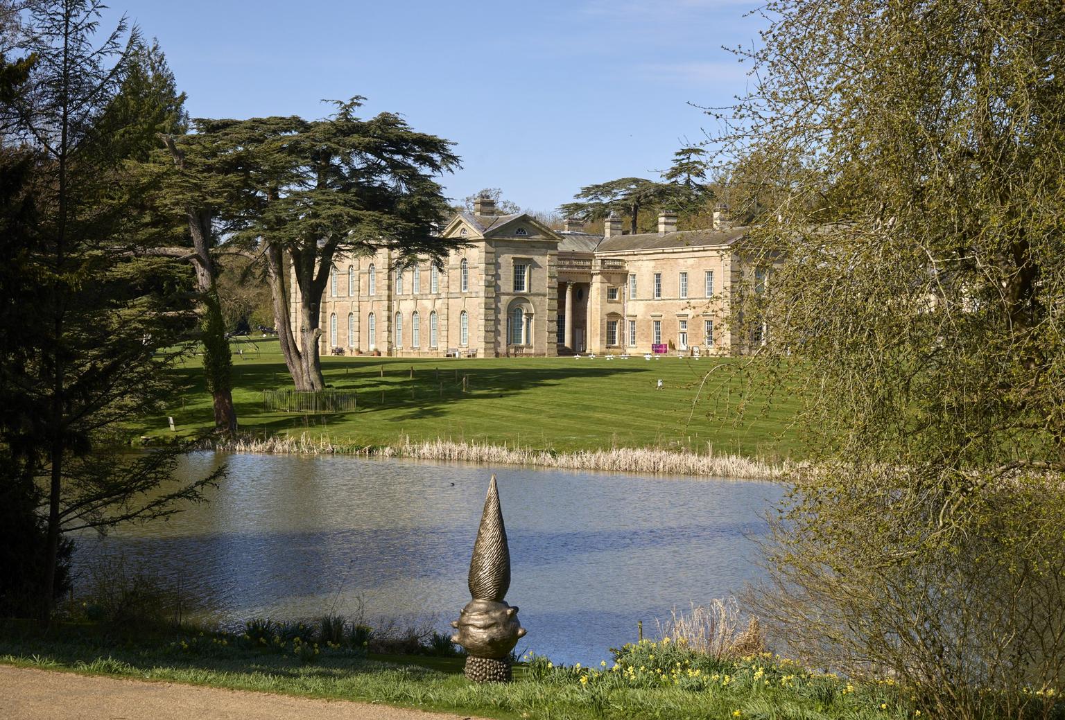 A beige bricked Georgian mansion overlooks a green lawn and a large lake. A metal pointy sculpture sits on the other side of the lake.