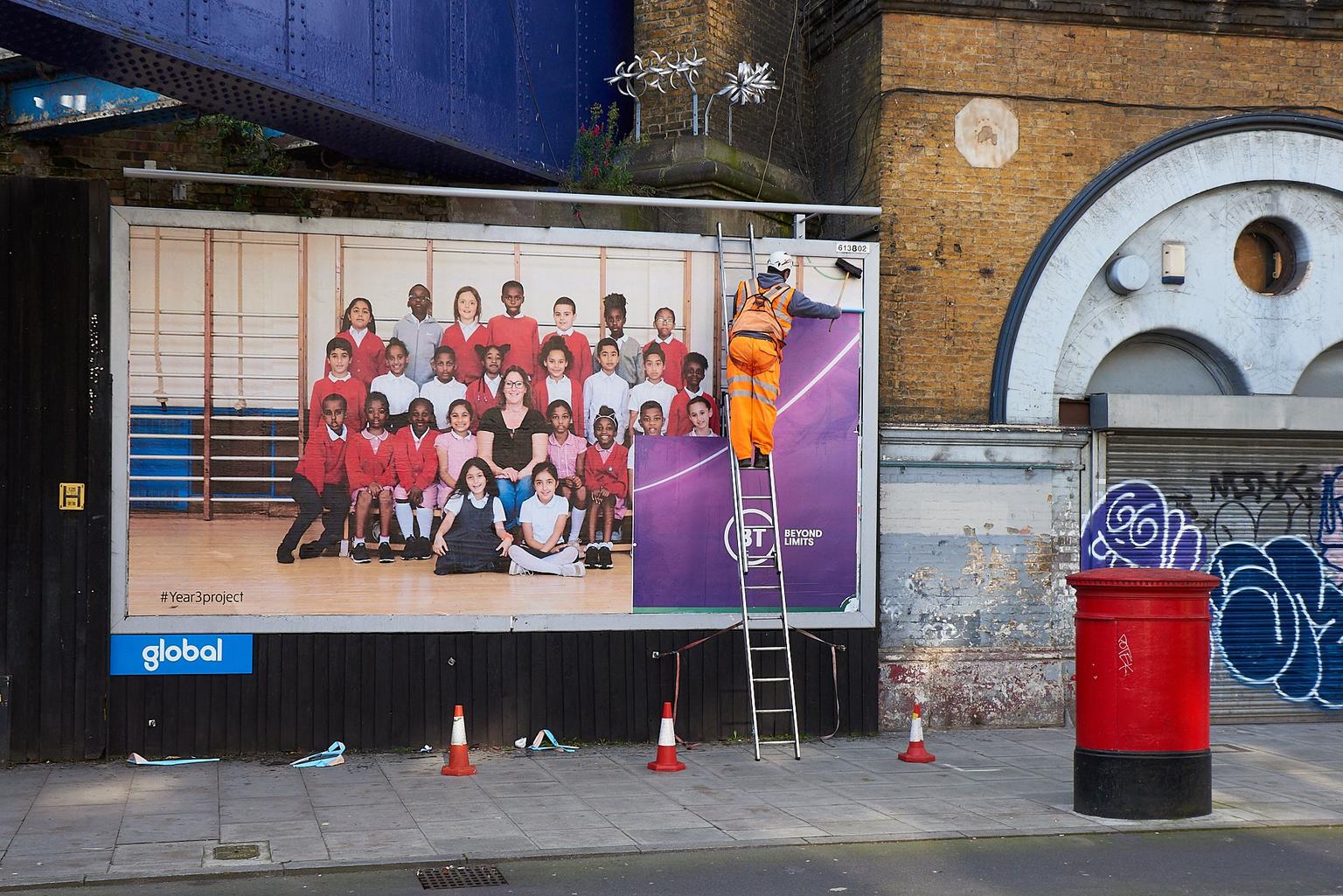 A billboard being installed by a man on a ladder wearing orange high-vis on a street of a photograph of a group of Year 7 school children in their uniform. About half are wearing red cardigans and jumpers; the other half are wearing white polo shirts.
