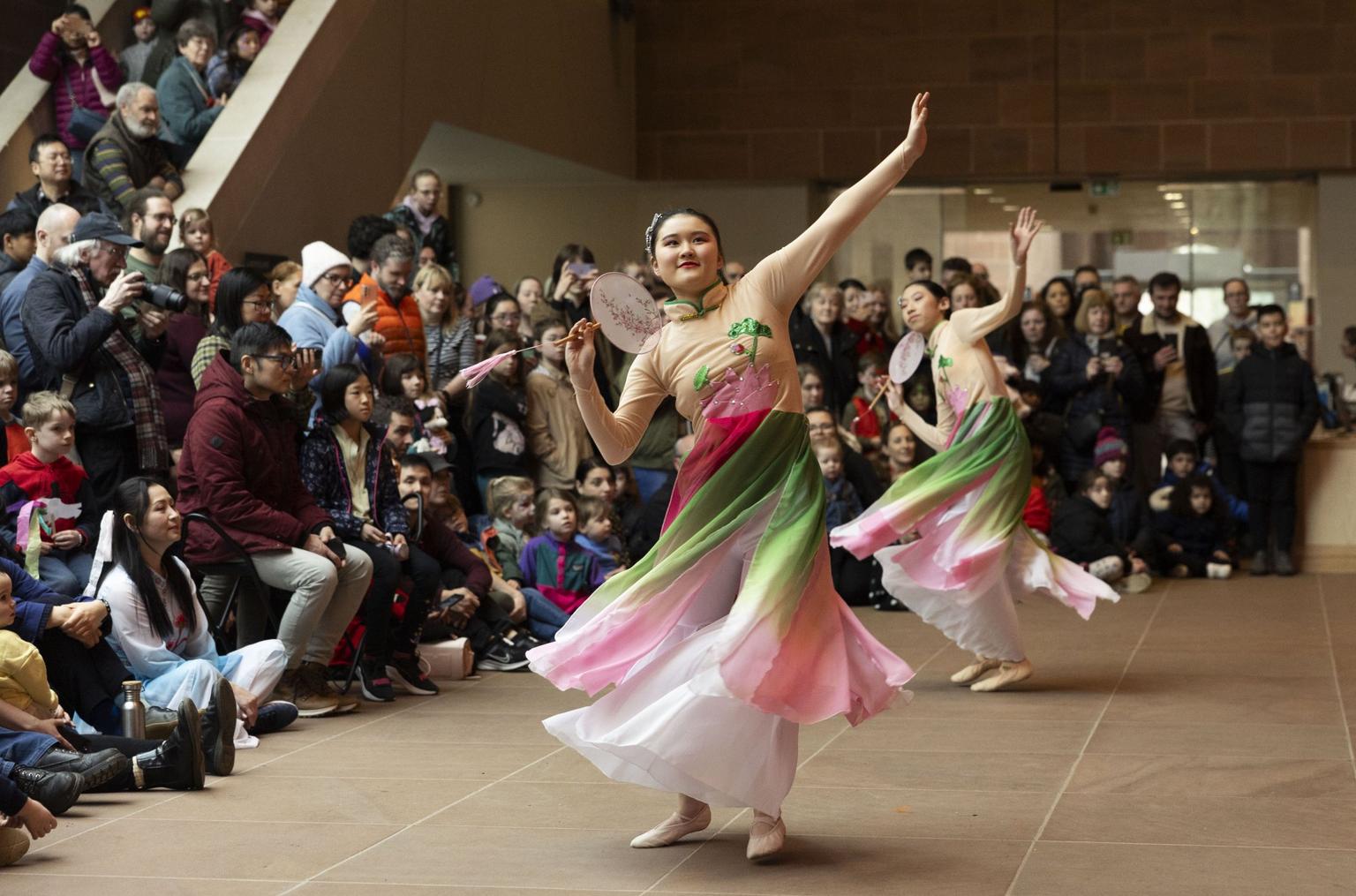 Two dancers in flowing pink and green dresses perform a graceful dance with hand-held fans before a seated crowd in an indoor public space.