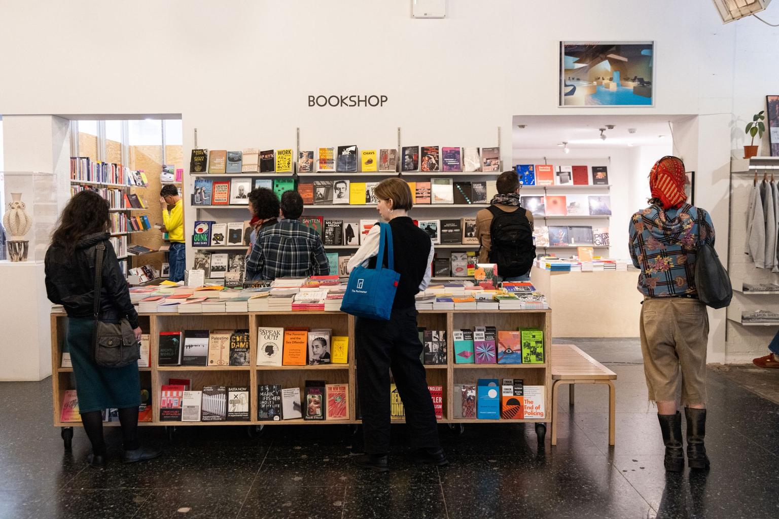 Several customers browse a white-walled bookshop with a central table of colorful books.