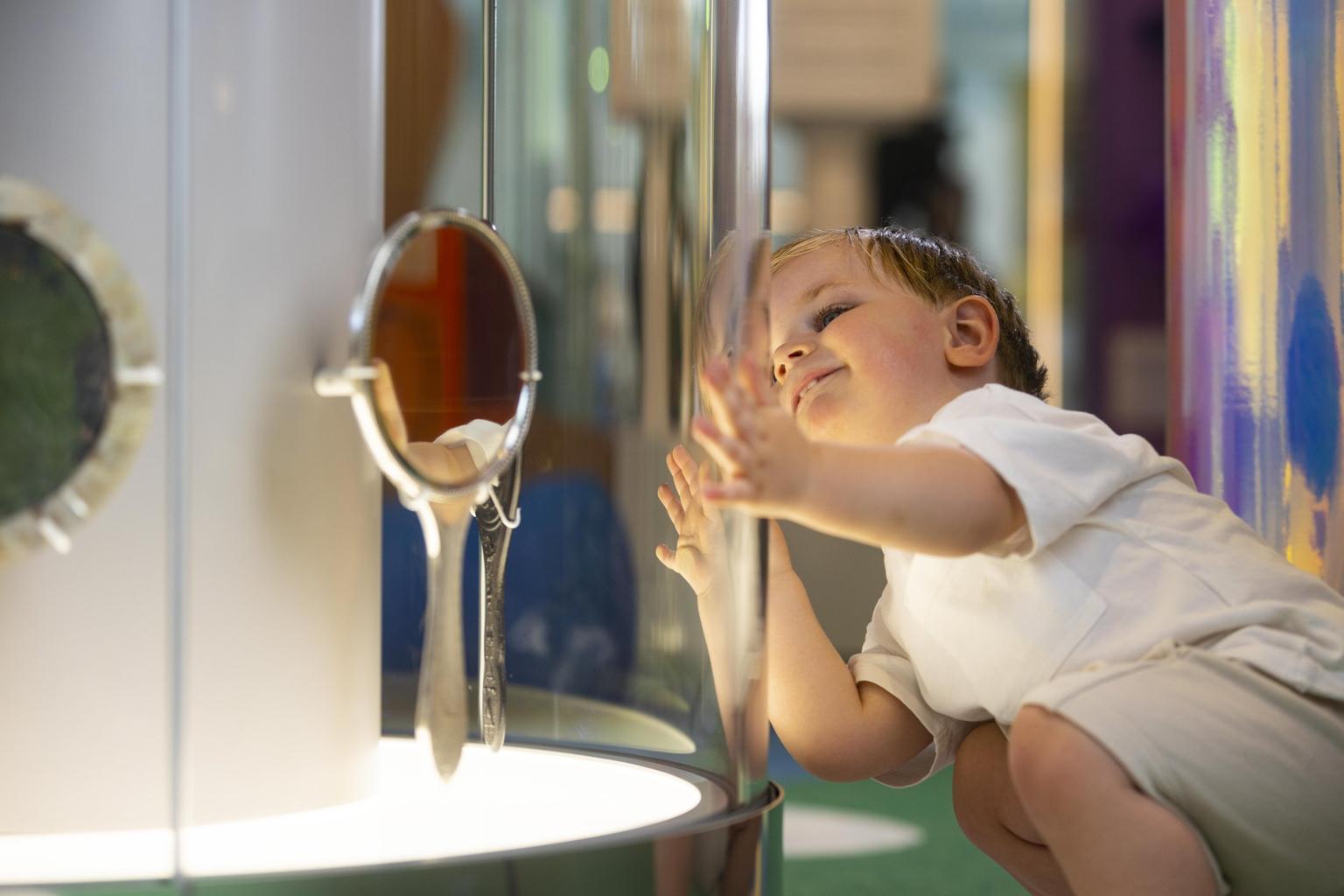 A young boy in a white shirt presses his hands against a clear exhibit panel while curiously peering at reflective objects inside.