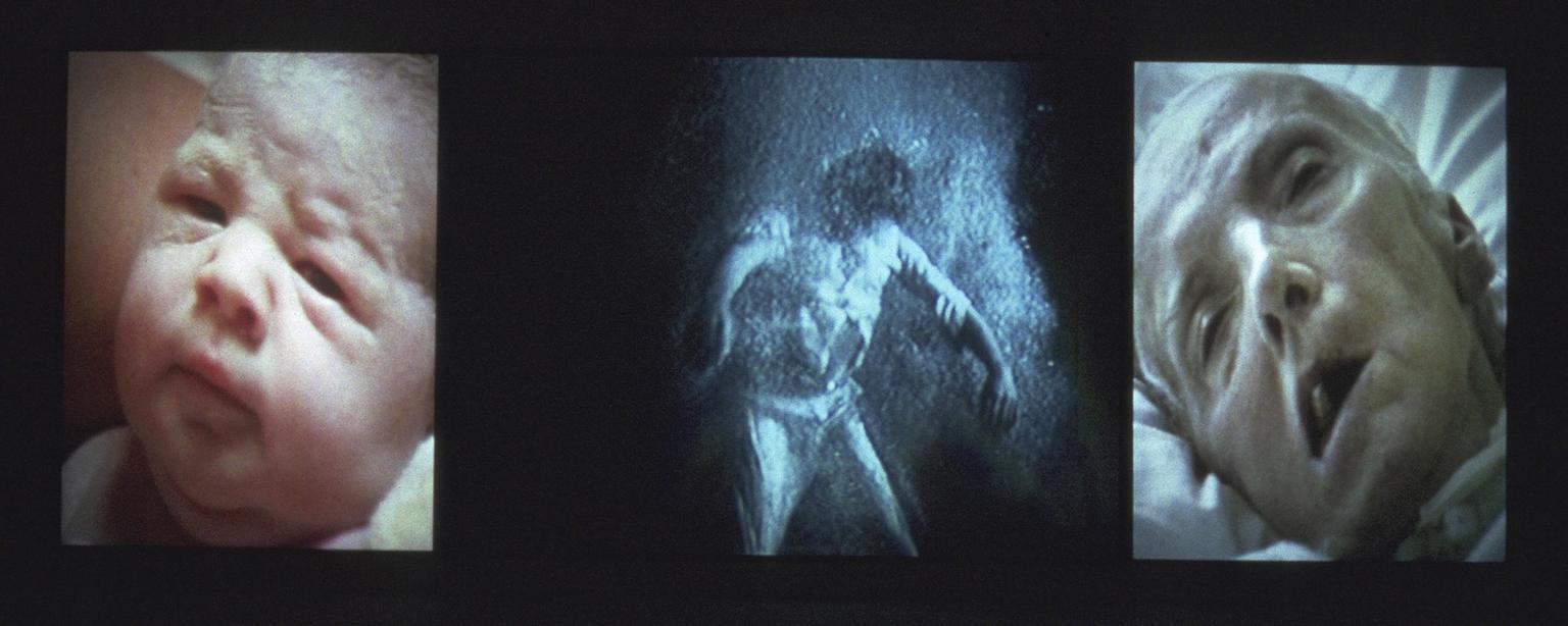 Three large-scale panels of projected video images form a configuration based on the tripych altarpiece. The left panel shows an image of a young woman in the process of giving birth. the right panel shows an image of an old woman in the process of dying. Both are documents of actual events. The central panel shows an image of a clothed man underwater moving through alternate stages of turbulence and undulating stillness, held in fragile suspension before an indistinct, shadowy space, suspended between birth and death.