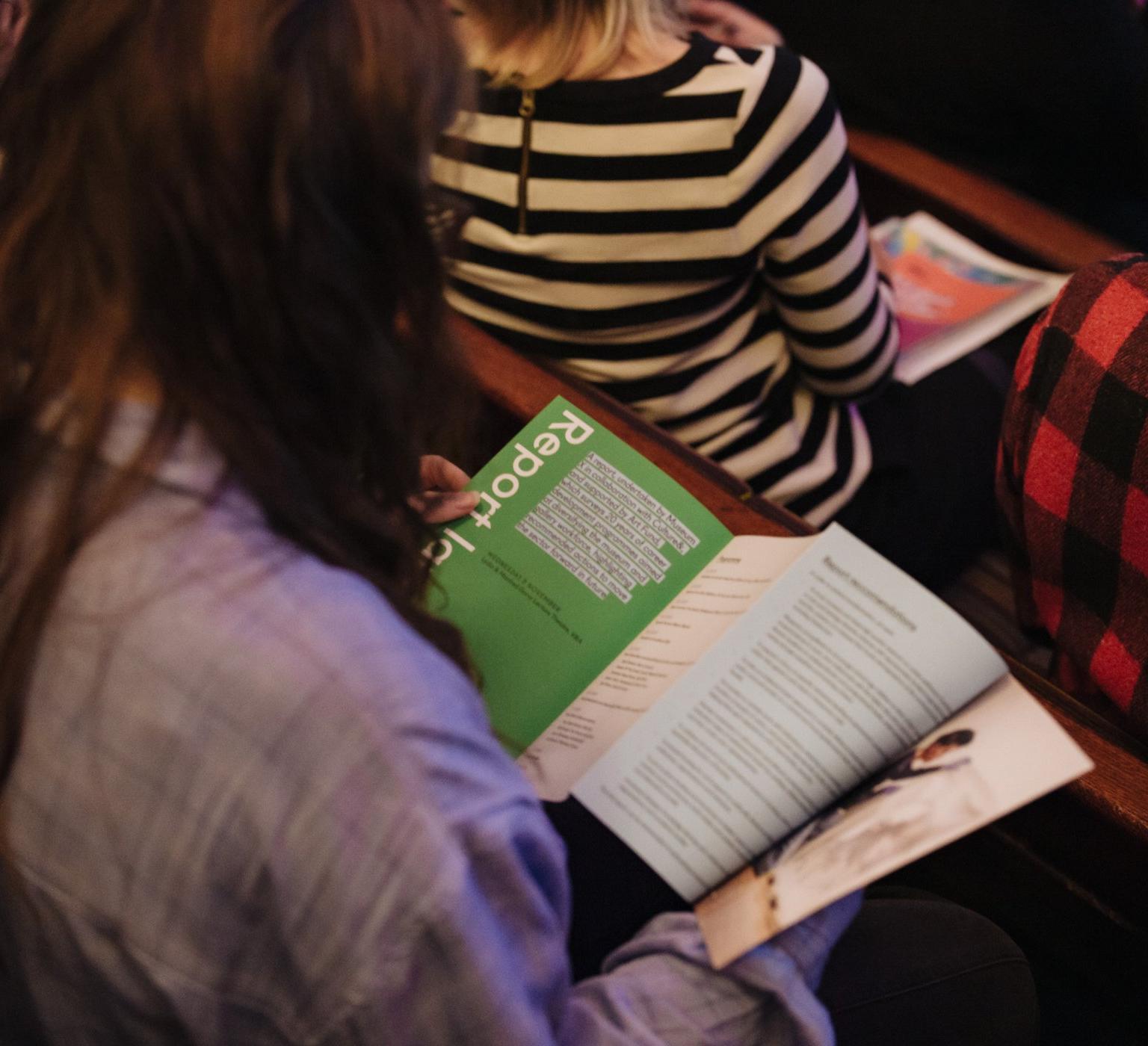 A person in a light shirt reads a green report booklet while seated among an audience.