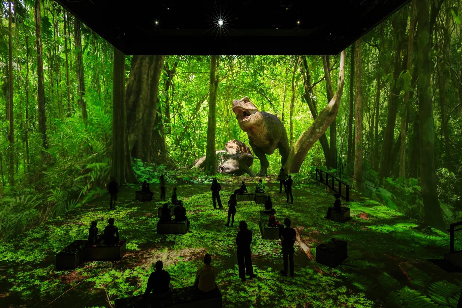 Visitors watch a giant roaring T. rex projected in a vivid green jungle in an immersive exhibition.