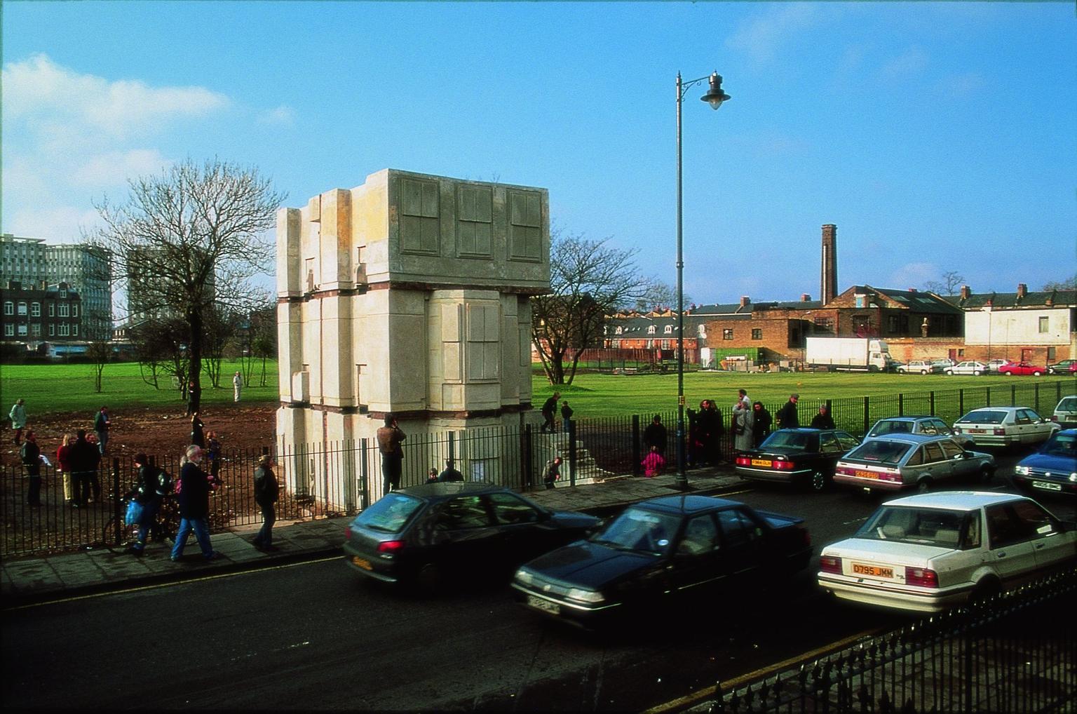 A life-size concrete model of a three storey-house on the edge of a park. Lots of people are walking around it, looking at it and taking pictures.