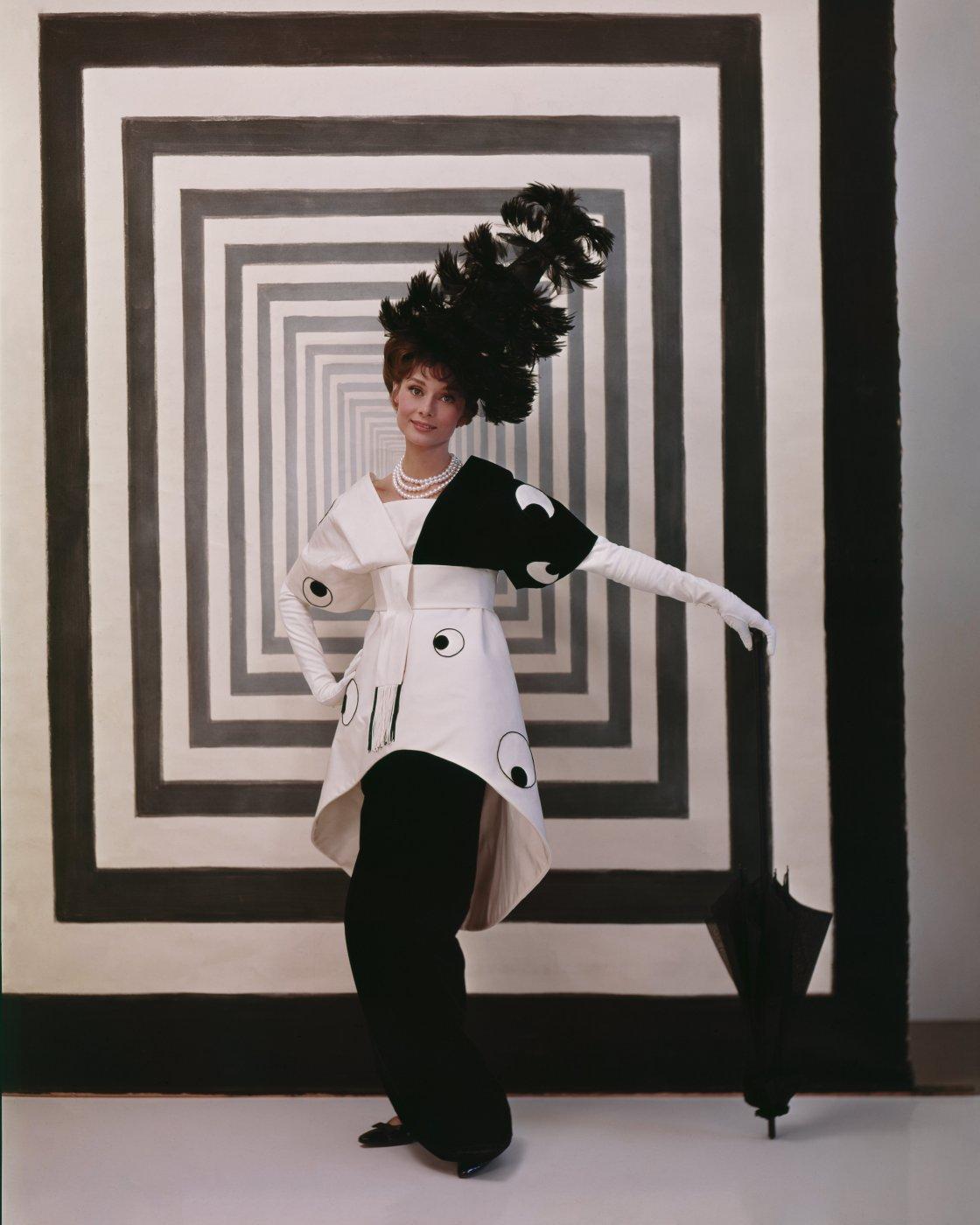 Audrey Hepburn in a black-and-white dress and feathered hat poses with a black umbrella in front of a concentric black-and-white square backdrop.