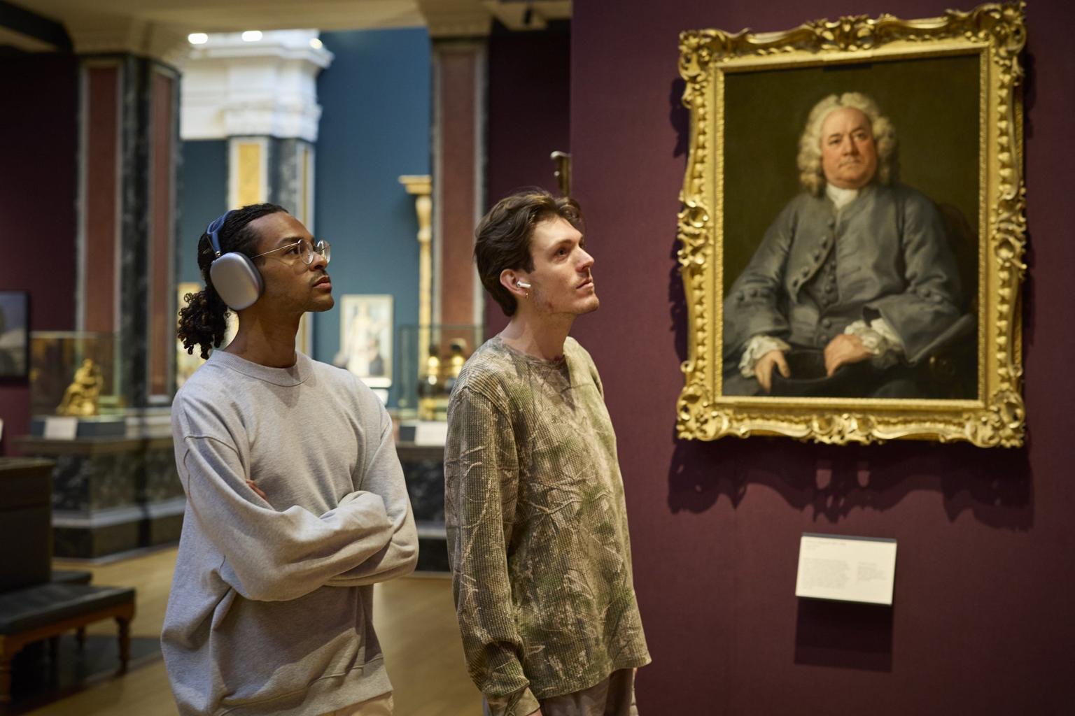 Two visitors stand in a museum gallery, one wearing headphones, both studying something out of frame. A large framed portrait hangs on a burgundy wall behind.
