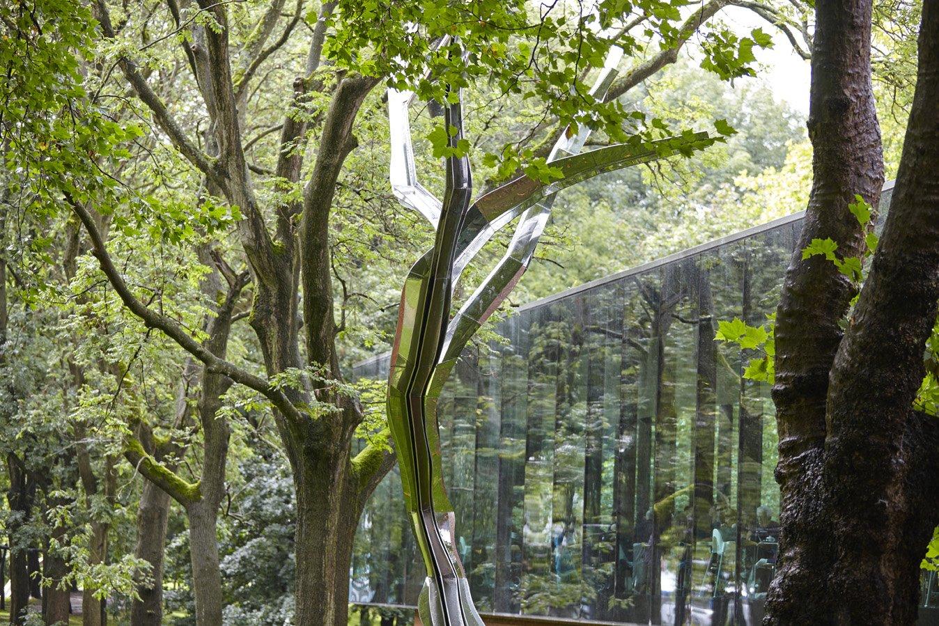 A silver sculpture of a tree surrounded by real trees and with a glass building beyond