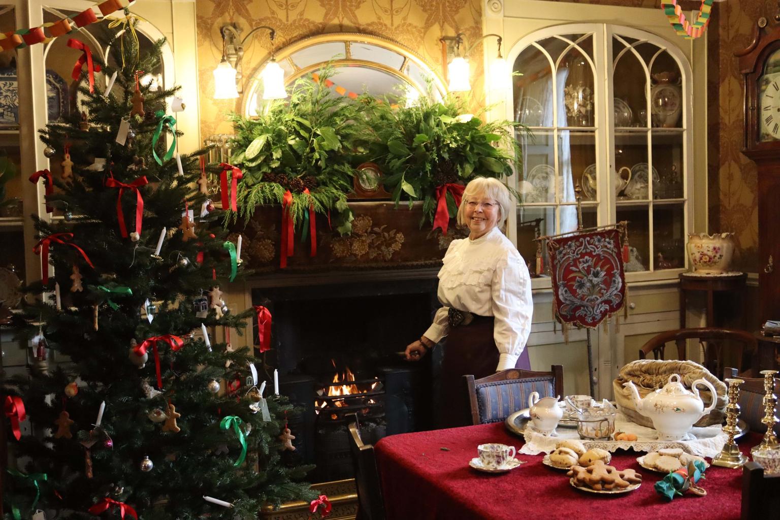 A woman in a white blouse smiles while tending a roaring fireplace in a decorated room with a Christmas tree and table of biscuits and tea.