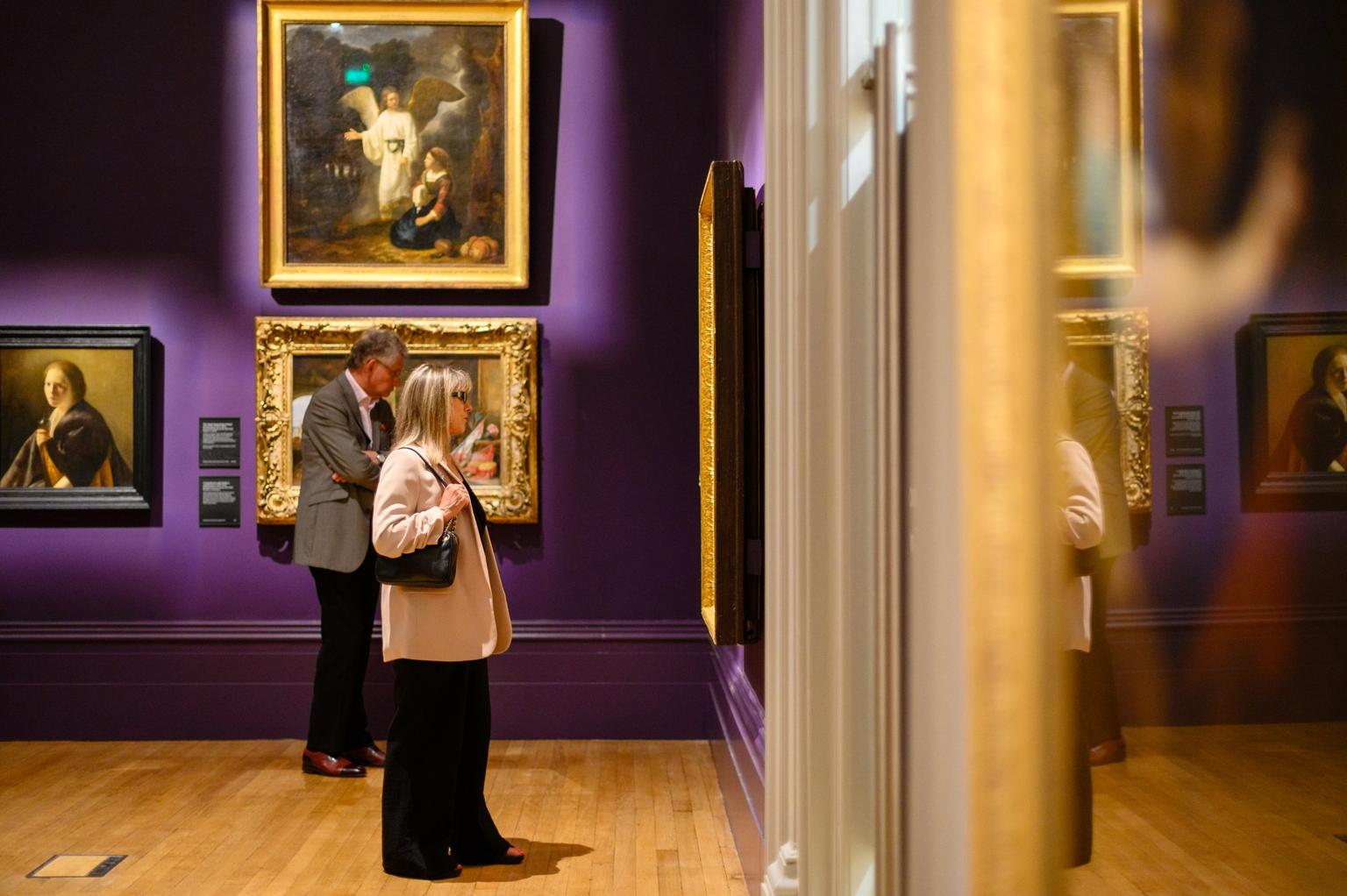 Two people looking at paintings in gold frames on a purple wall