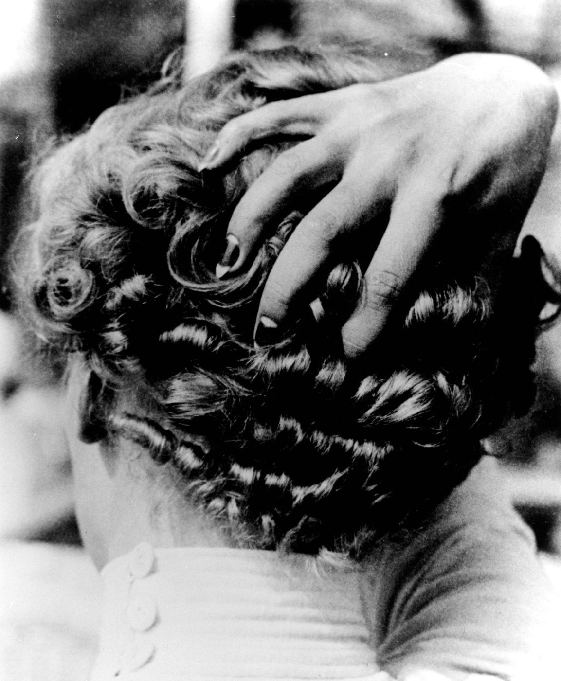 Black-and-white photo of a person seen from behind with a hand resting on short, curly hair.