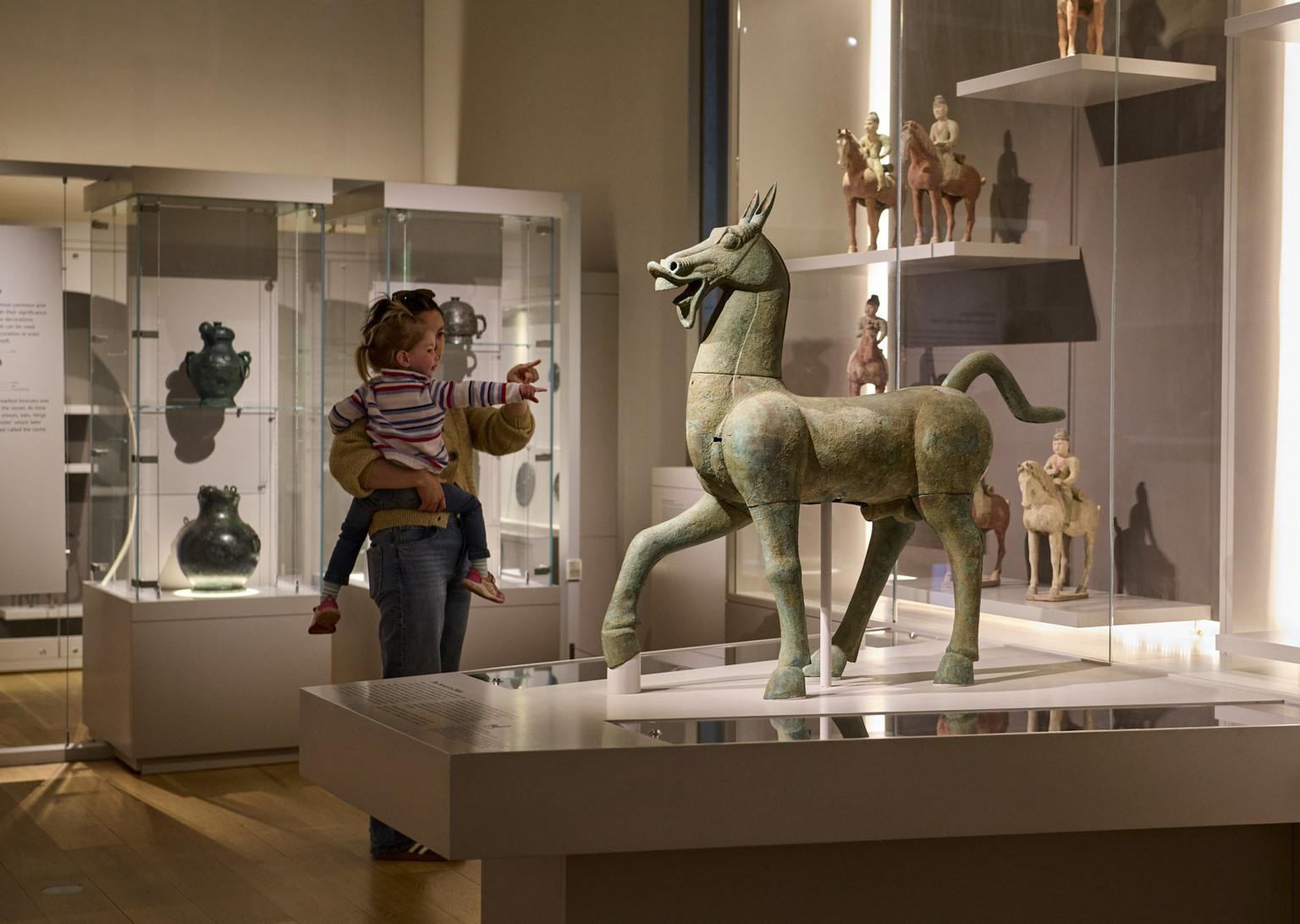 A woman carries a small child in a gallery space displaying ancient Chinese ceramic horses.