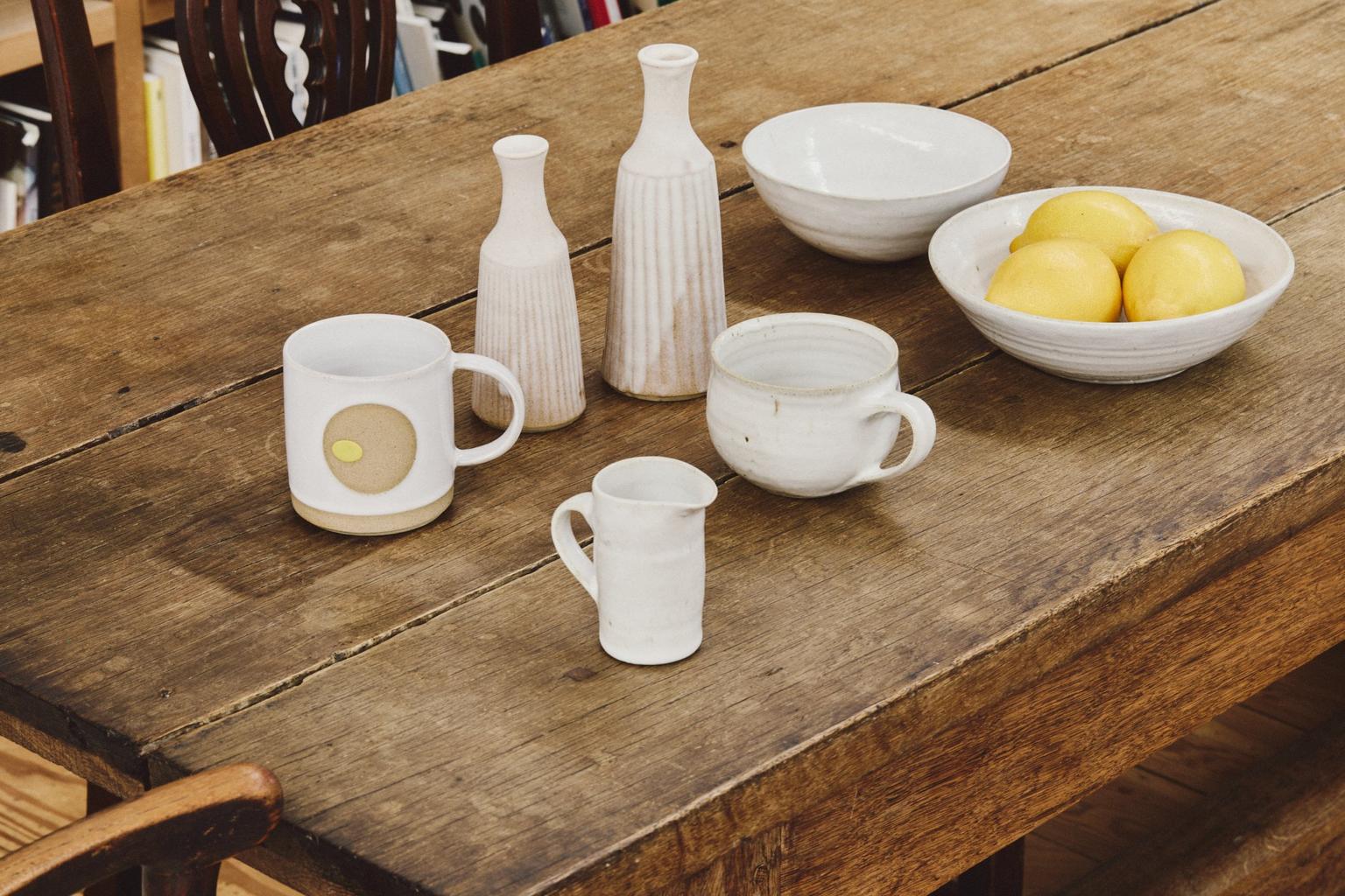 A rustic wooden table holds white ceramic vases, mugs, and a bowl of lemons.