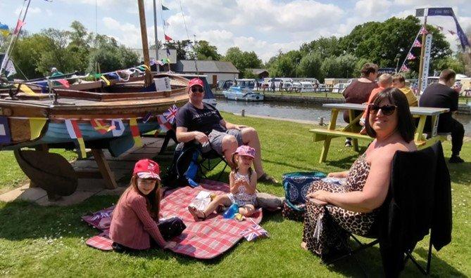 A family sit around a picnic blanket in front of a small boat by a river