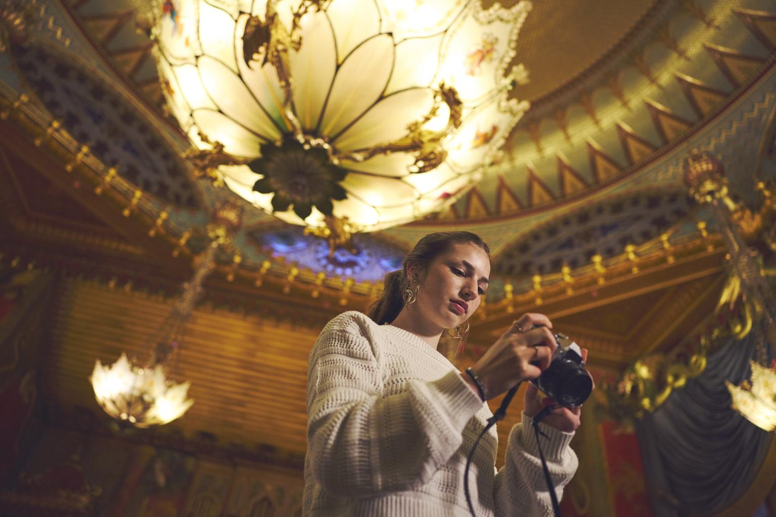 A woman in a white sweater adjusts a camera beneath ornate golden chandeliers and an elaborate ceiling.