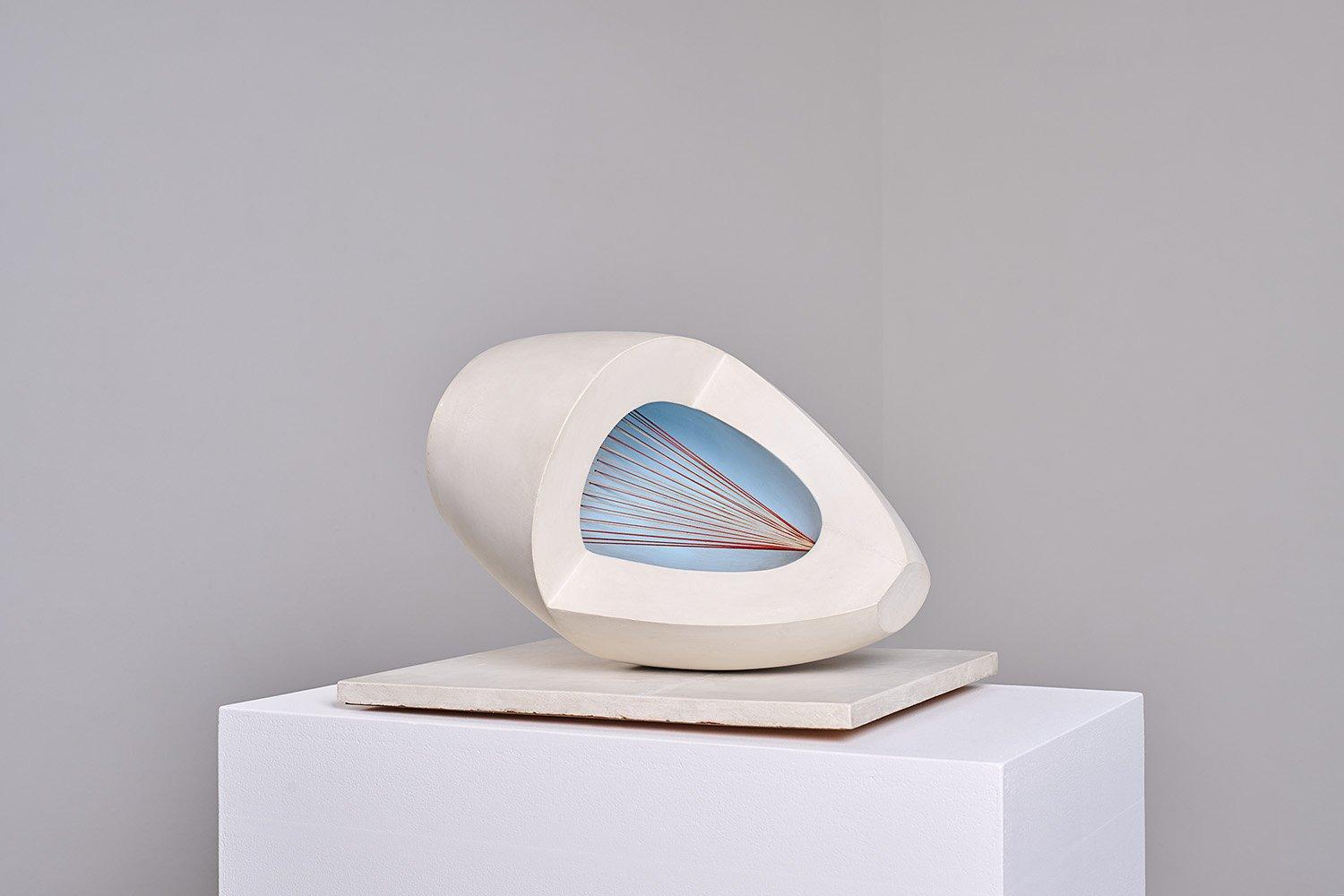 A modern abstract sculpture resembling a hollowed, egg-shaped form, partially sliced open to reveal an interior painted sky blue. Inside, a series of taut red strings radiate from a single point at the base toward the open edge, creating a geometric fan-like pattern. The sculpture rests on a white plinth against a neutral grey background.