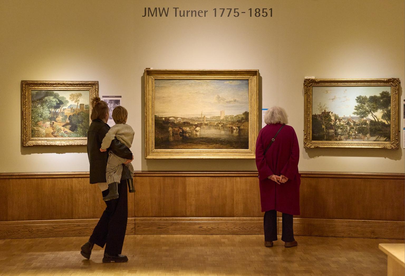 Two museum visitors, one holding a child, stand before landscape paintings by Turner in gilded frames.