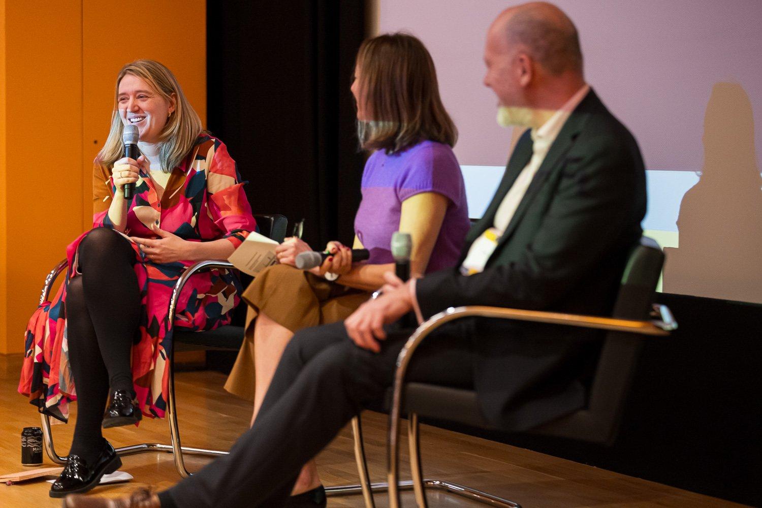 A smiling woman in a colourful pink and red patterned dress speaks into a microphone during a panel discussion on stage.