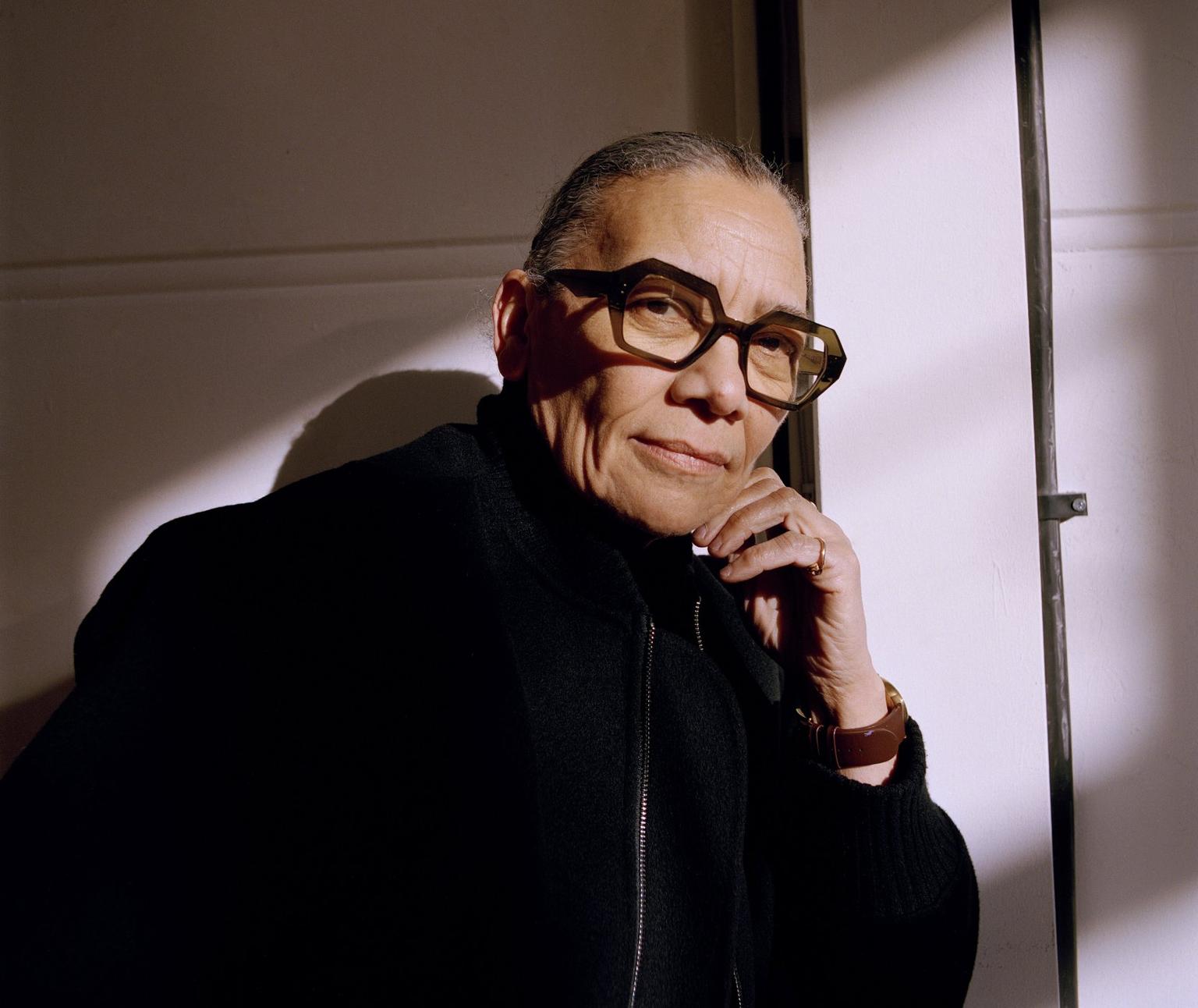 The artist Lubaina Himid resting her hand on her chin in warm side lighting against a pale wall.
