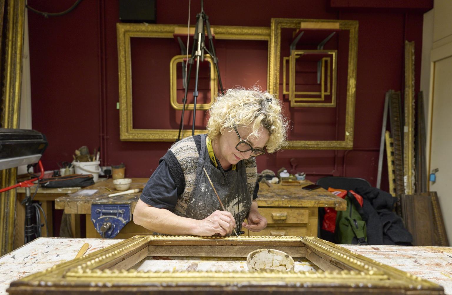 A conservator carefully restores a gilded picture frame with a small brush in a workshop with red walls and stacked gold frames.