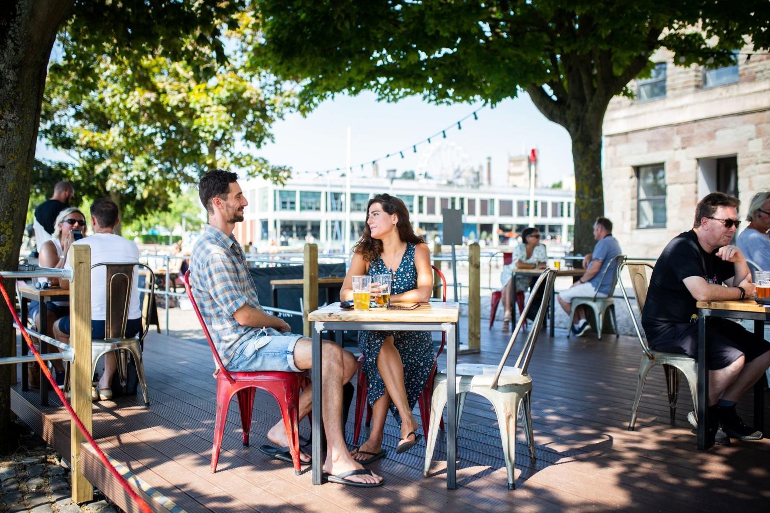 A man and a woman sitting on a sunny outdoor terrace at a table, drinking and chatting; groups of other people are sat at other tables around them.