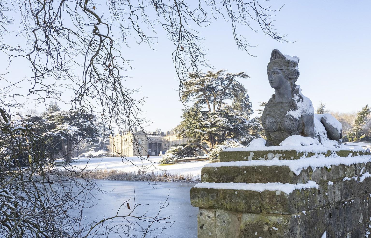 A snow covered Sphinx statue in the foreground, snowy grounds and a stately house behind