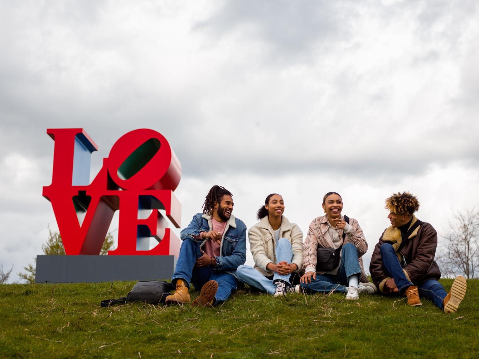 A group of for people sitting on a grassy hill with a sculpture spelling LOVE behind them