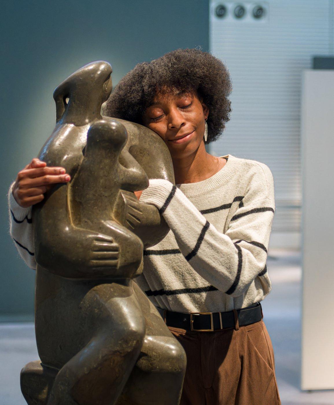 A person in a striped sweater gently hugs a smooth dark stone sculpture with eyes closed in a quiet gallery setting.