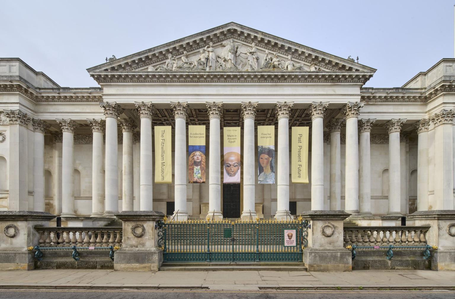 Neoclassical museum facade with tall columns, sculpted pediment and vertical exhibition banners overlook a green and gold gate.