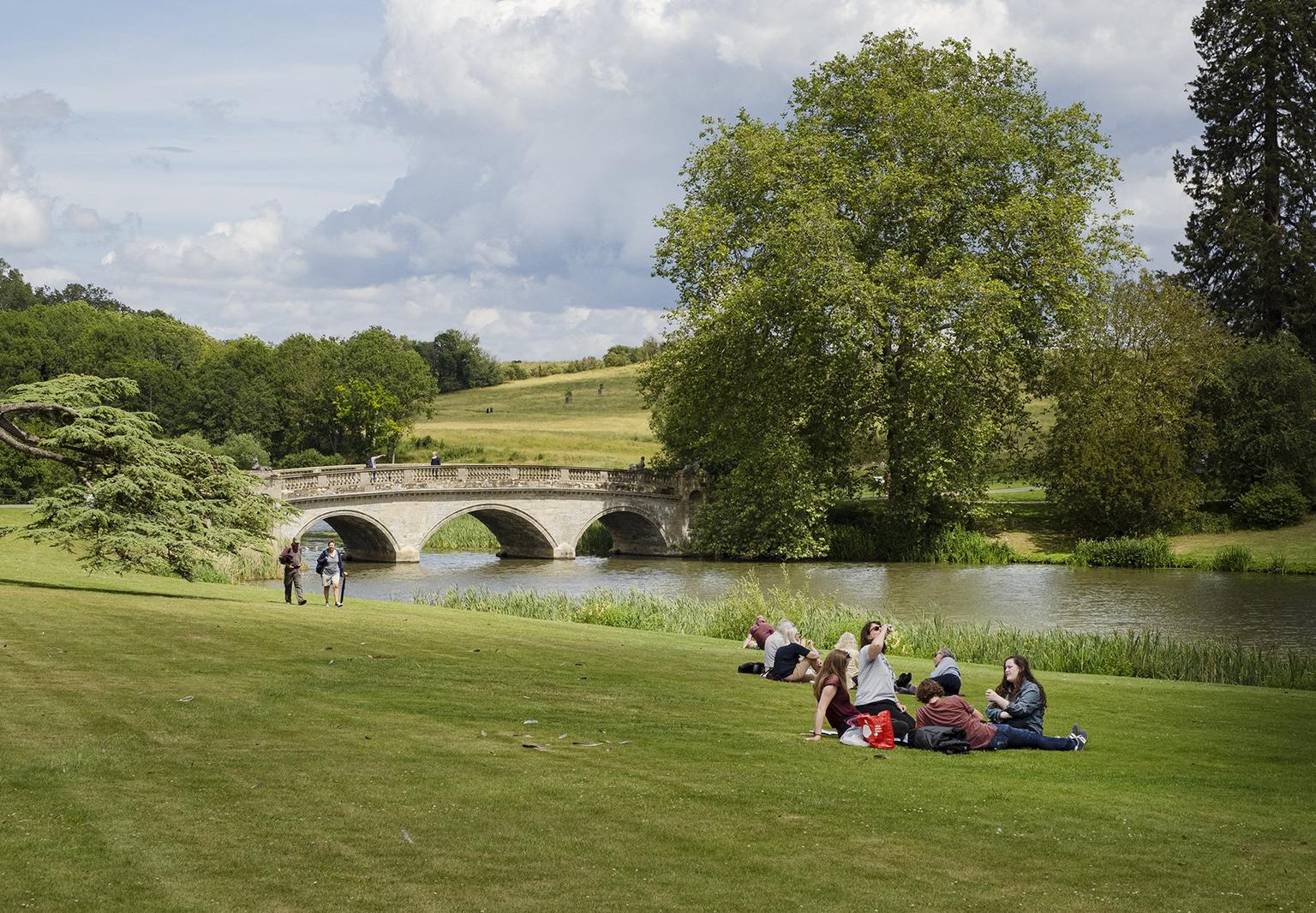 A stone bridge over a lake, people sitting in groups on the grassy banks