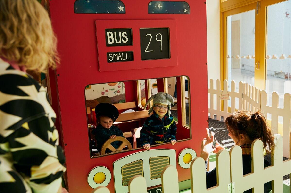 Two children play in a child-sized red double decker bus as their parents watch on.