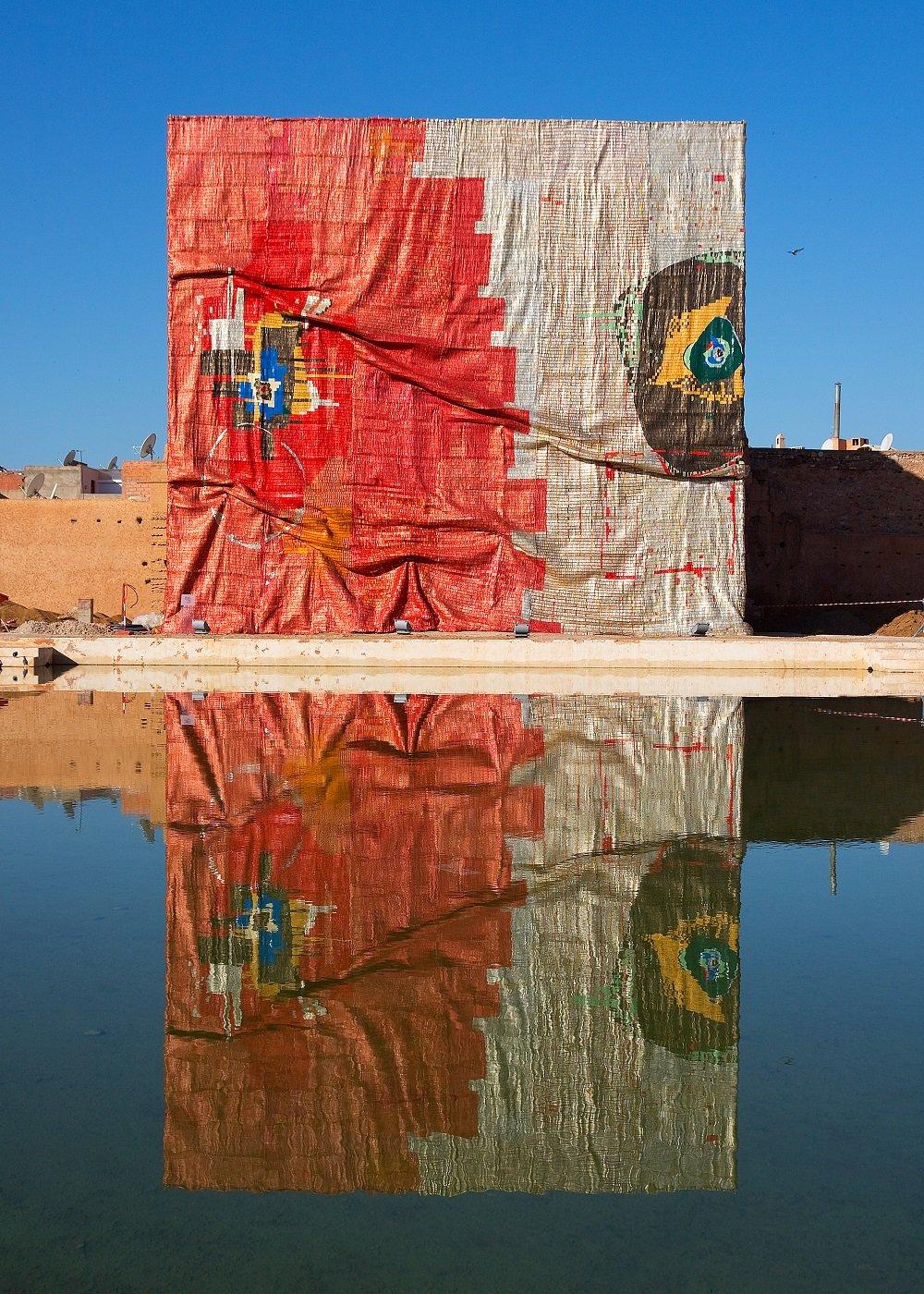 Monumental mostly red- and silver-coloured aluminium work, covering the entire facade of a building and reflected in a body of still water.