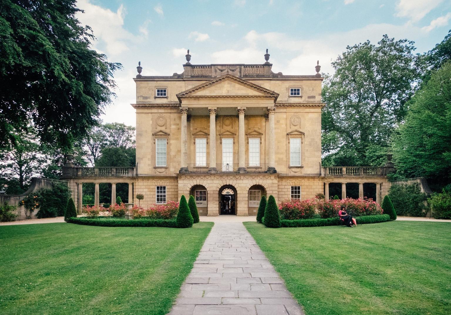 A grand, sand-coloured, three storey Georgian house in a neoclassical style stands in front of a manicured lawn with symmetrically pruned hedges and bushes with pink flowers.