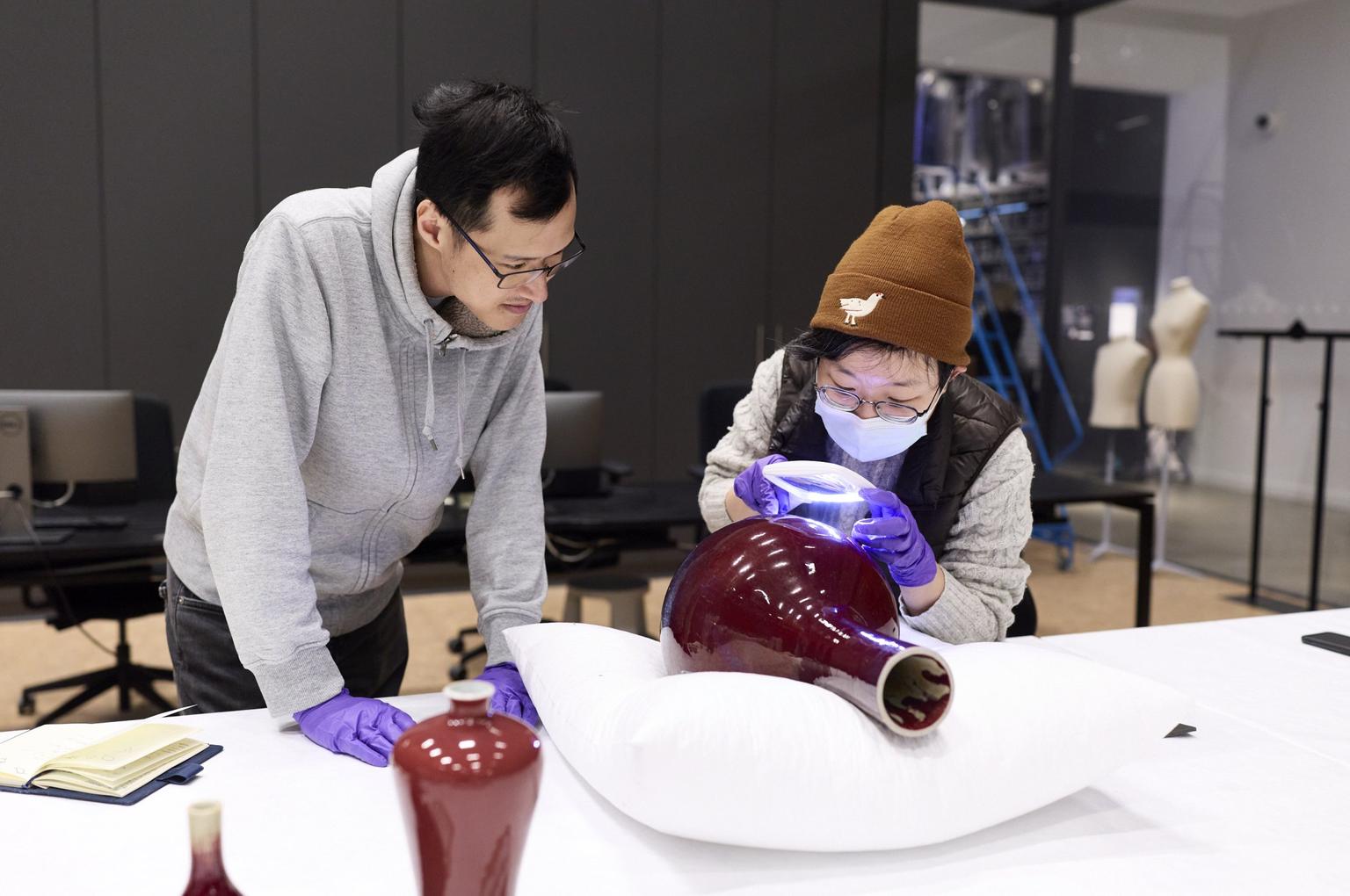 Two people wearing purple latex gloves examine a red ceramic vase; one is using a handheld light while the other leans in.