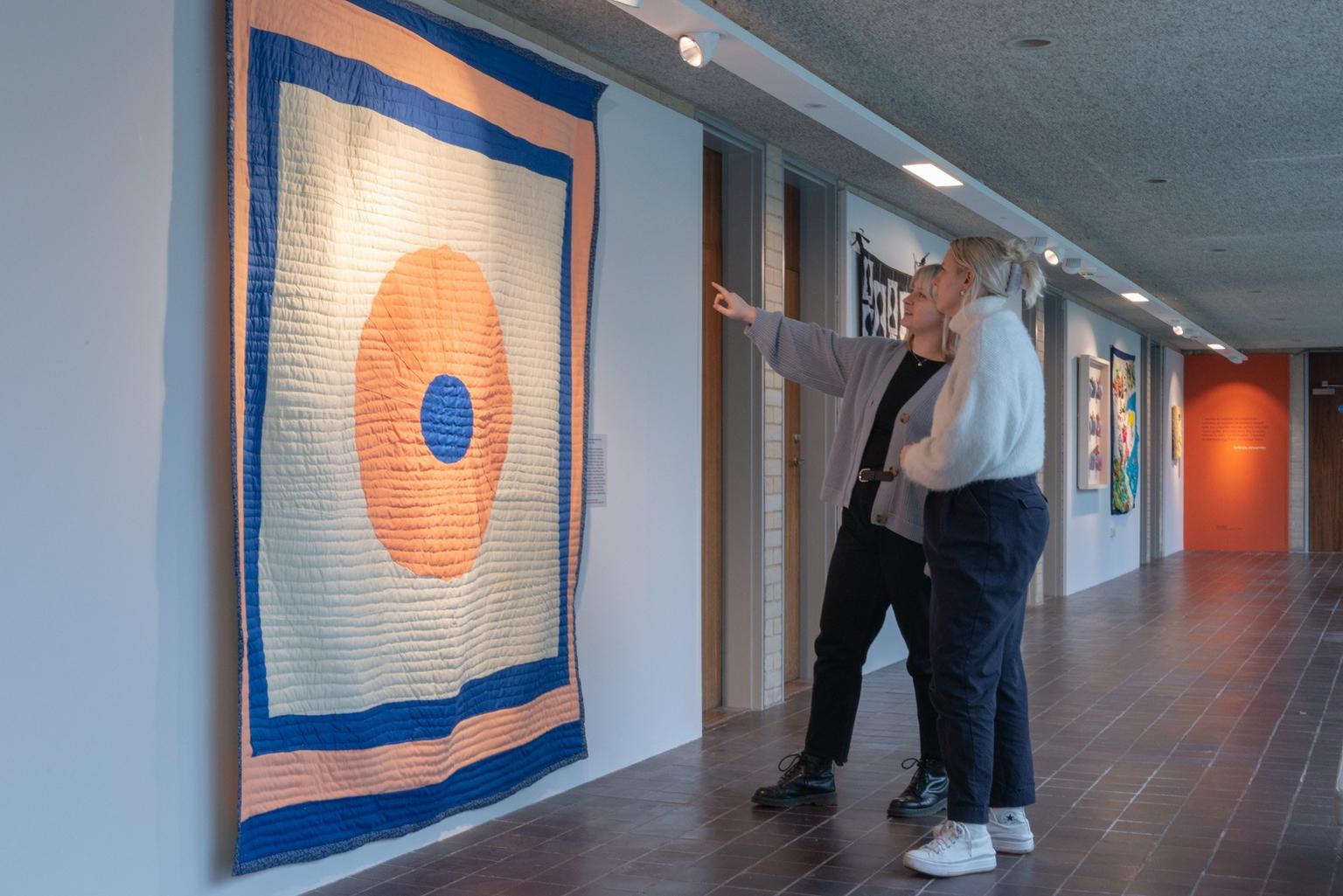 Two women standing inside an art gallery; one is pointing at a large blue and orange abtract textile hanging on the floor while they both look at it.