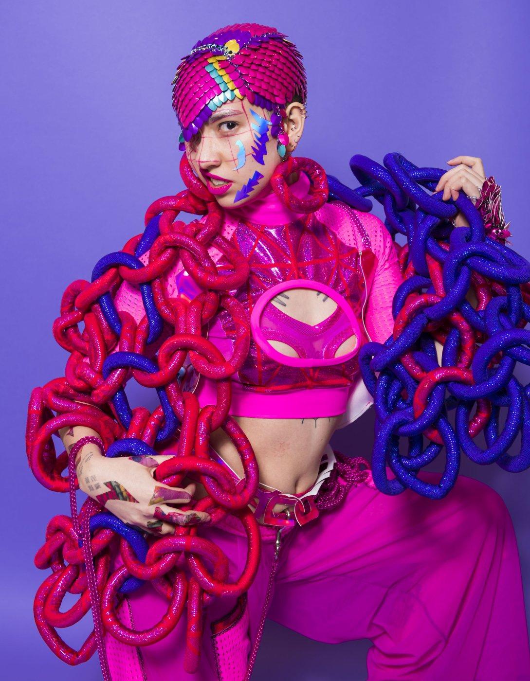 A person wears hot pink trousers with a matching top, a pink head cap and a pink and blue sculptural shroud. Blue metallic patterns and pink lines are drawn on their face.