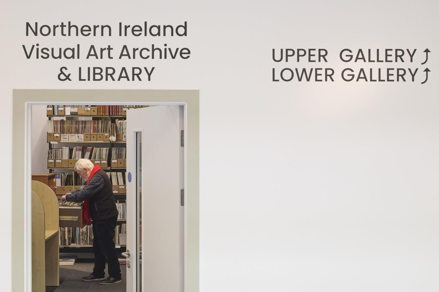 A white wall above a door with a sign that reads 'Norther Ireland Visual Art Archive & Library'. Through the door, a man is looking through a draw of archival materials.