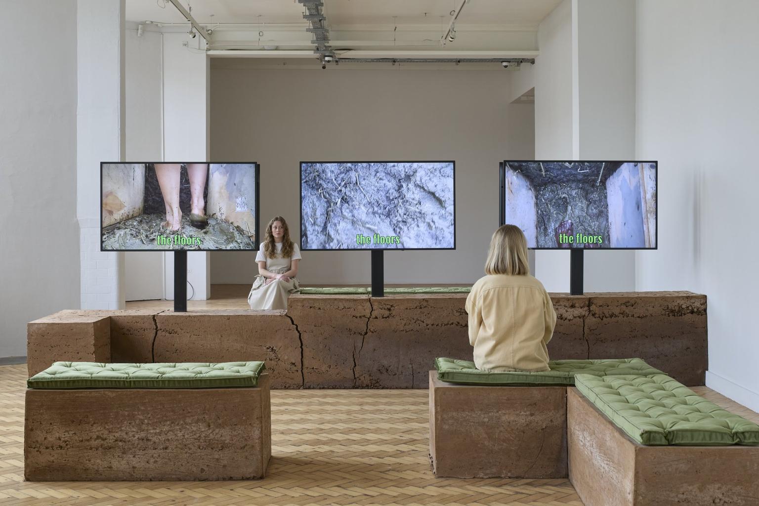 In a white-walled art gallery space, two women sit on rusty brown concrete blocks with green seating pads, watching three flat TV screens showing different shots from a film with 'the floors' overlaid in green text.