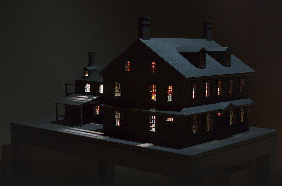 A dark miniature two-story house model sits on a pedestal with warm multicolored lights glowing through its windows.