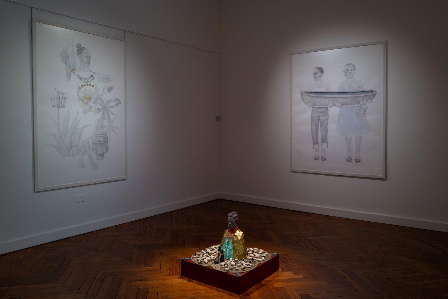 A small golden and turquoise figure sits on a low red plinth surrounded by shell-like objects under a spotlight in a dim gallery corner.