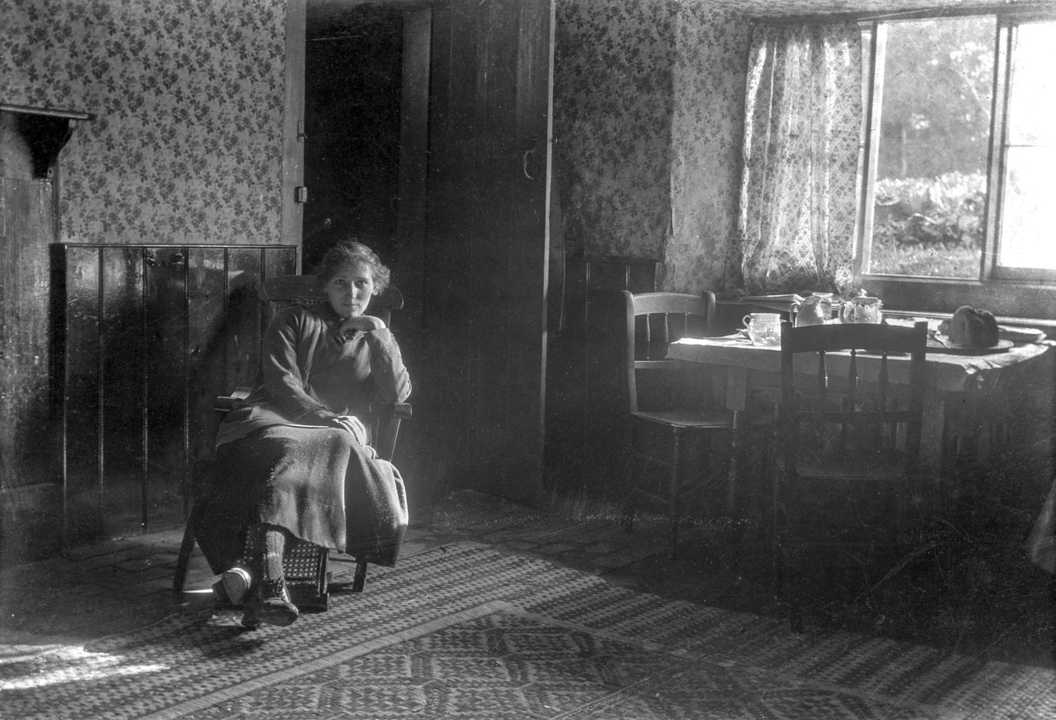 A black and white photograph of a woman sitting in a wooden chair in room decorated with floral wallpaper. To her left and in front of an open window is a table and chairs with a tea set.